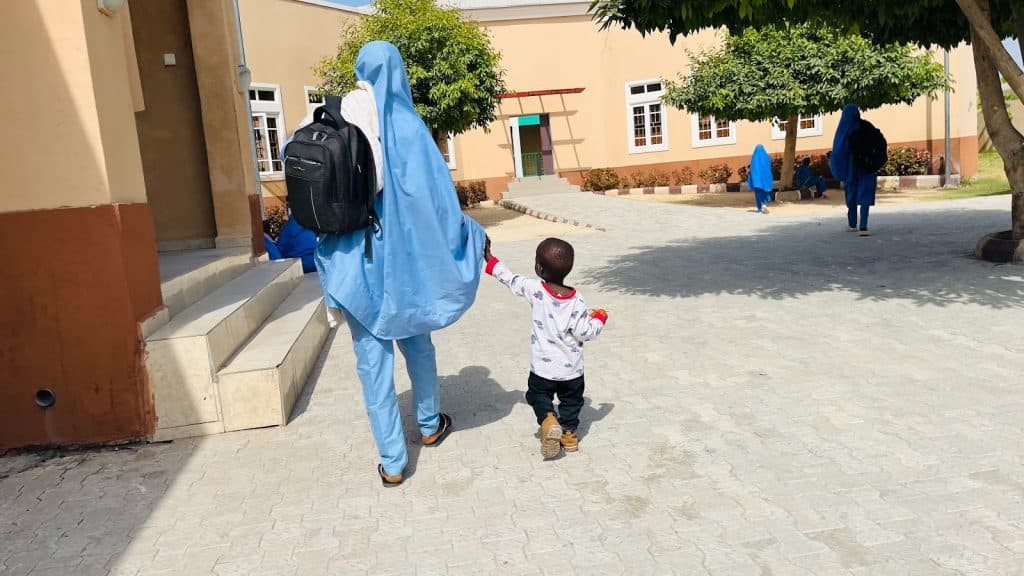 Child holding hands with a person in a blue outfit, walking along a sunny path near a building with trees.