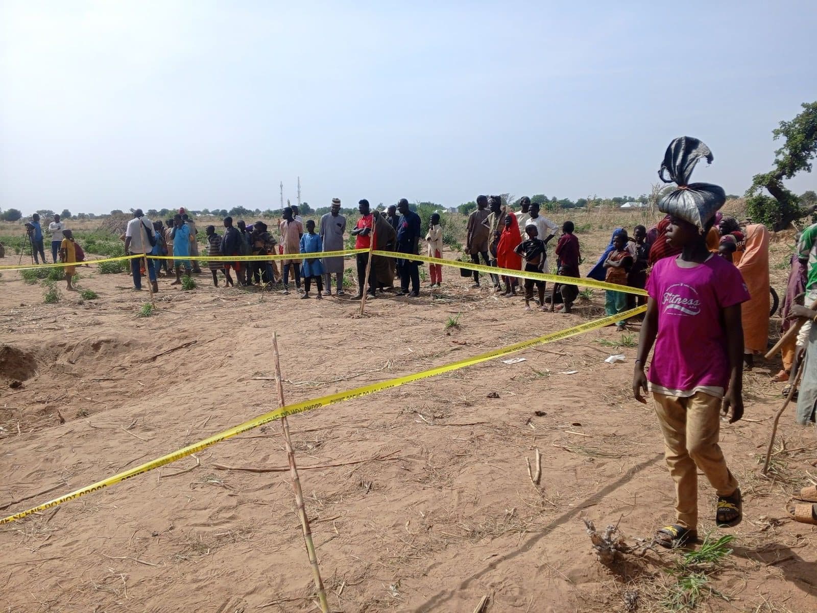 A group of people stands behind yellow caution tape on a sandy field under a clear sky, with sparse vegetation in the background.
