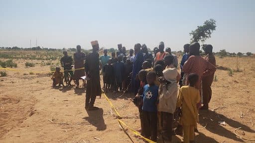 A group of people, including children, gather outdoors behind a yellow tape on a sunny day. Sparse vegetation is visible in the background.