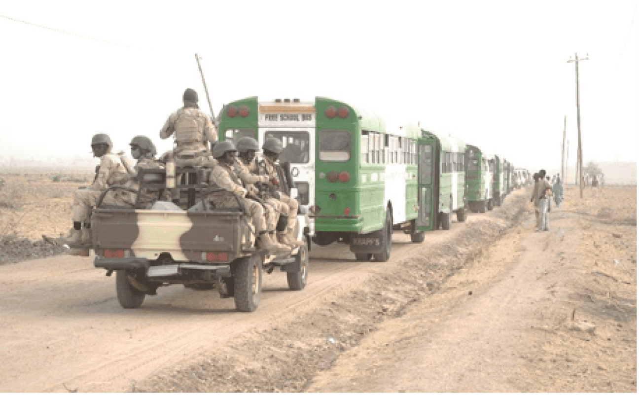 Military personnel on a jeep escort a convoy of green buses along a dirt road in a rural area.