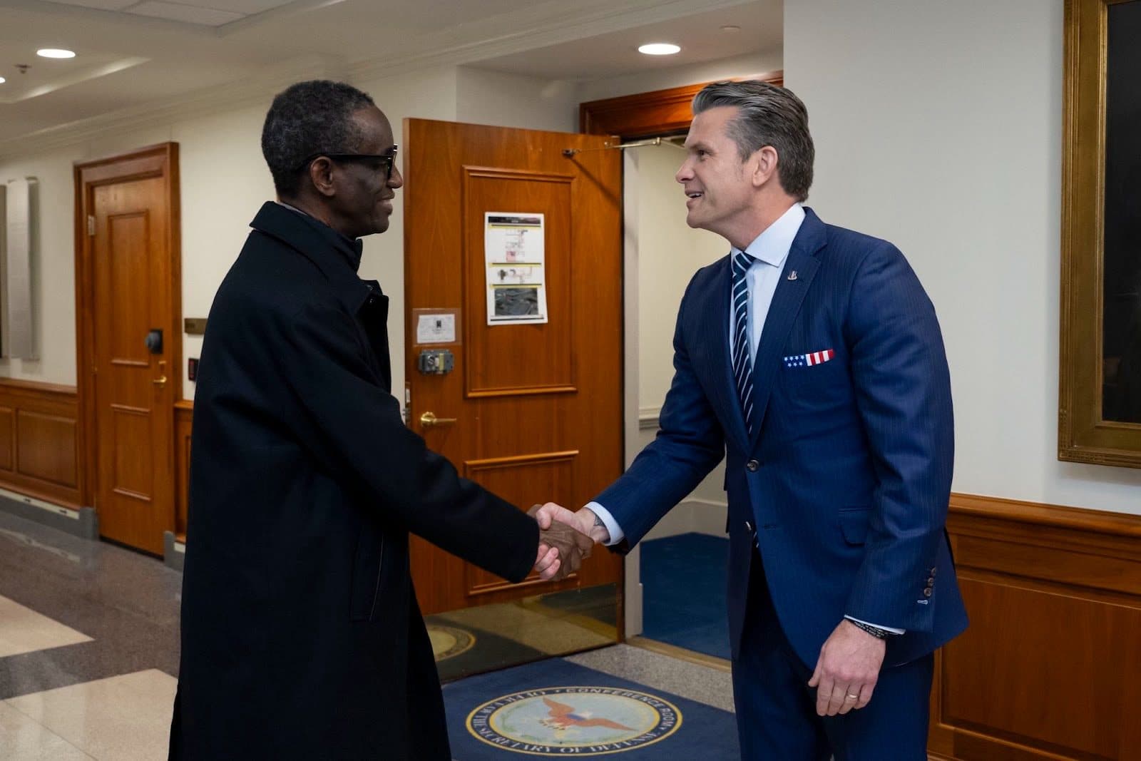 Two men in formal attire shaking hands in a hallway with wood paneling.