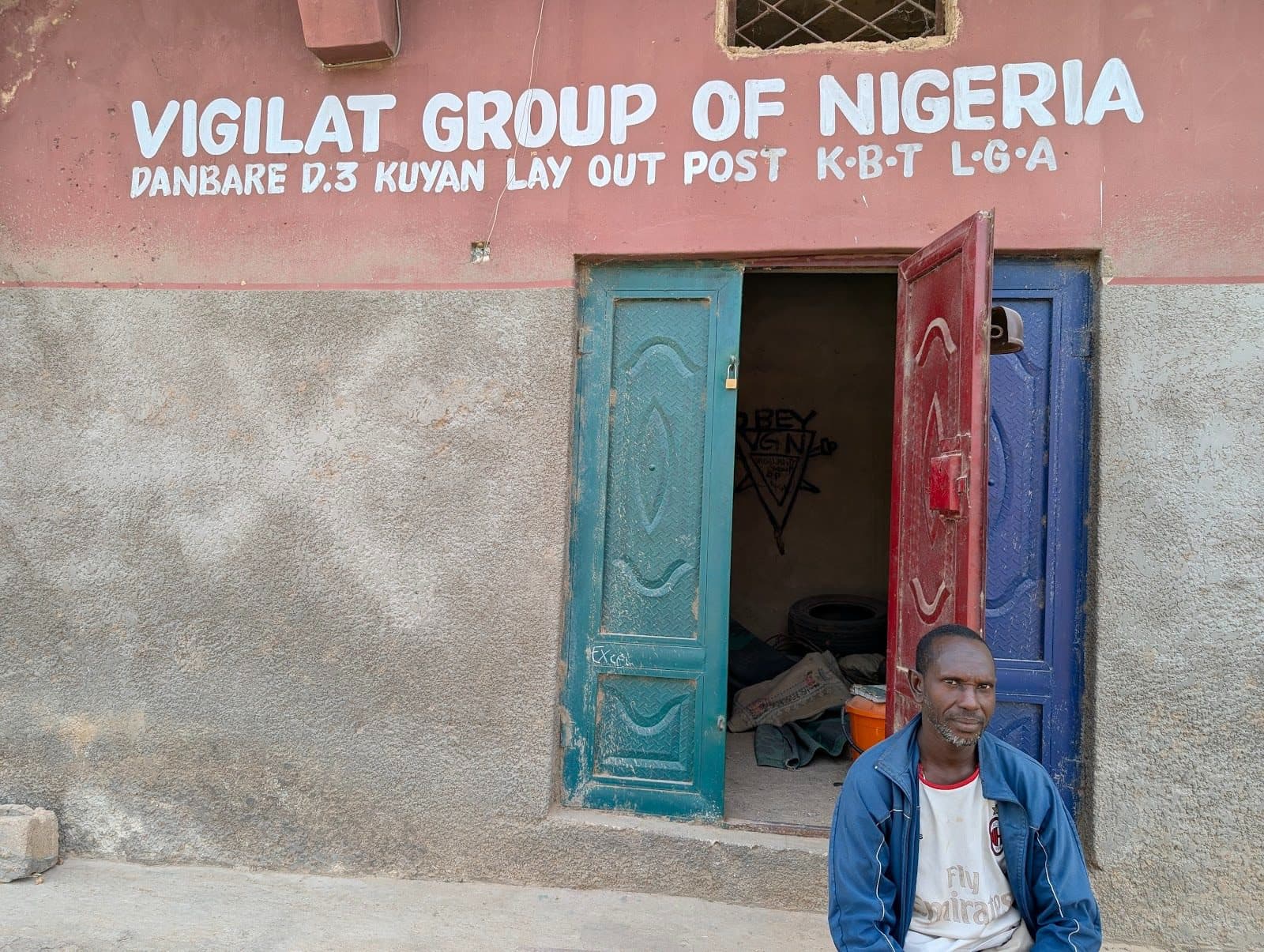 Man sitting outside a building with a sign reading "Vigilat Group of Nigeria" above blue and red open doors.