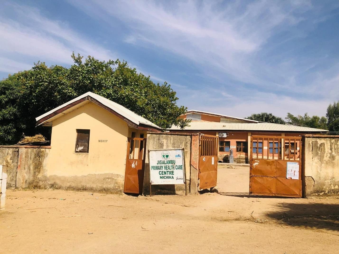 A rural health center with open gates, surrounded by trees under a blue sky.
