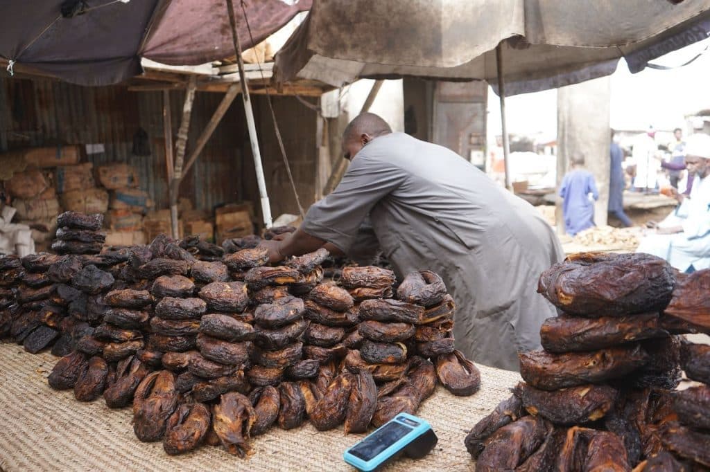 Man arranging smoked goods at an outdoor market stall, with a phone placed on the mat beside stacks of the product.