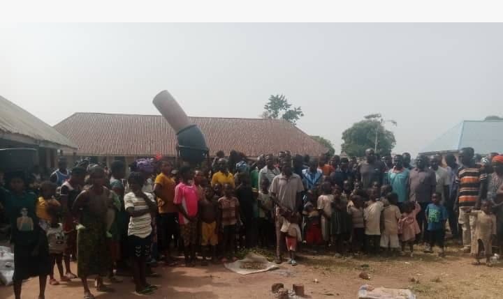 A large group of people, including children, standing outdoors near buildings in a rural setting.