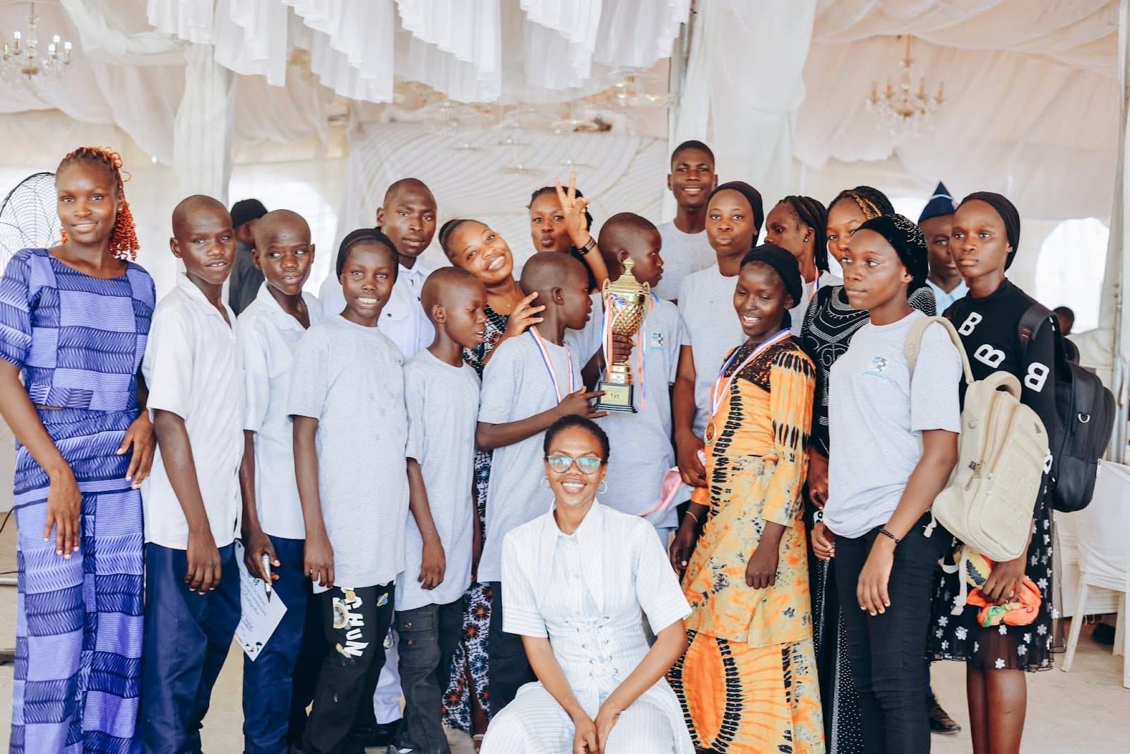 A group of people smiling, holding a trophy, indoors. Some wear matching gray shirts, others colorful attire, with a white draped backdrop.