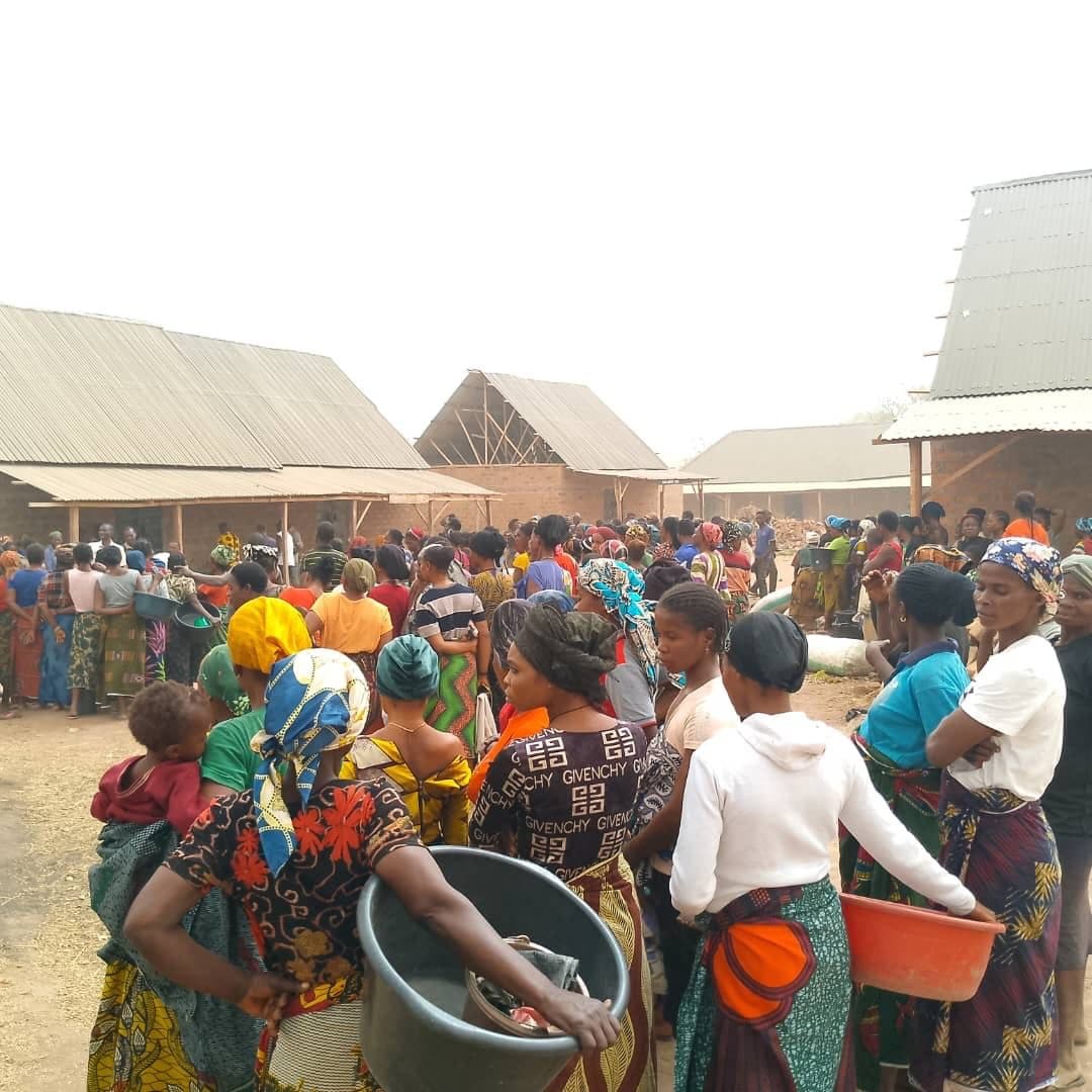 A large group of people gathered outside brick buildings with corrugated metal roofs, many holding containers and wearing colorful clothing.
