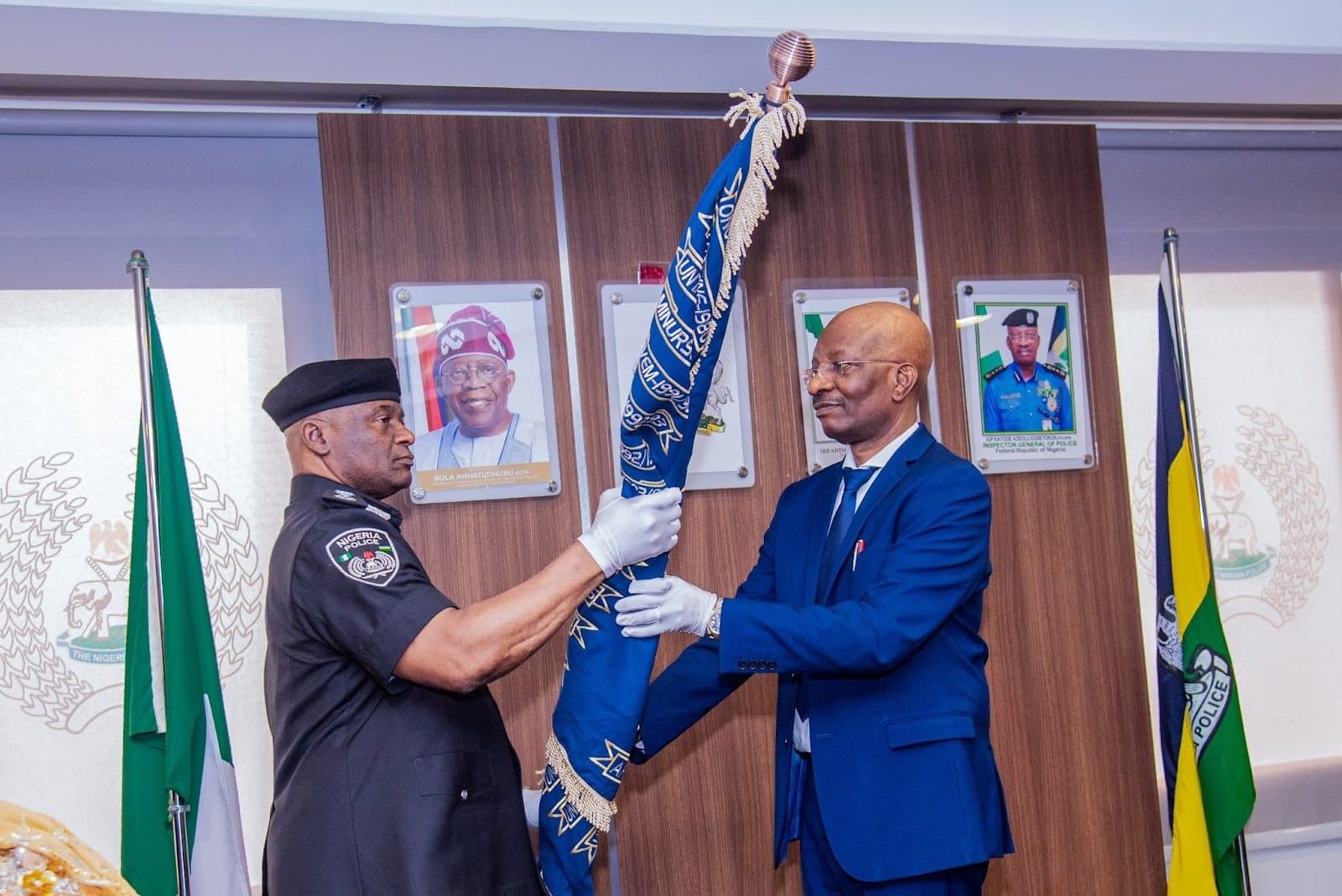 Two men exchange a decorated staff in an official setting, with flags and portraits in the background.