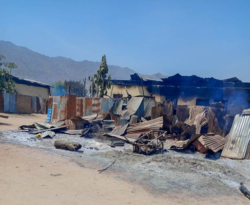 Corrugated metal structures burned and collapsed against a mountainous backdrop under a clear blue sky.