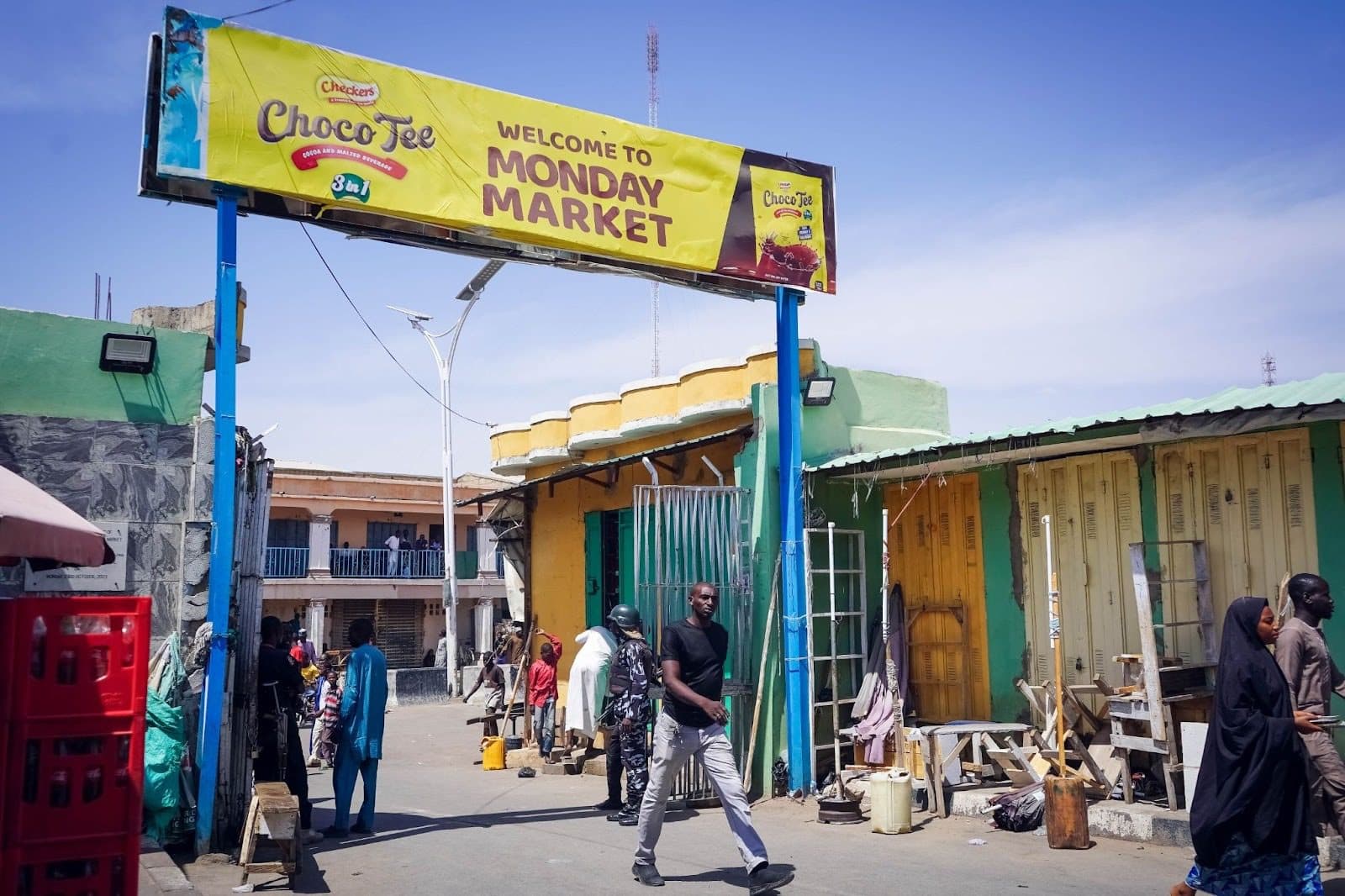 People walk under a "Welcome to Monday Market" sign in an outdoor market with colorful buildings and stalls.