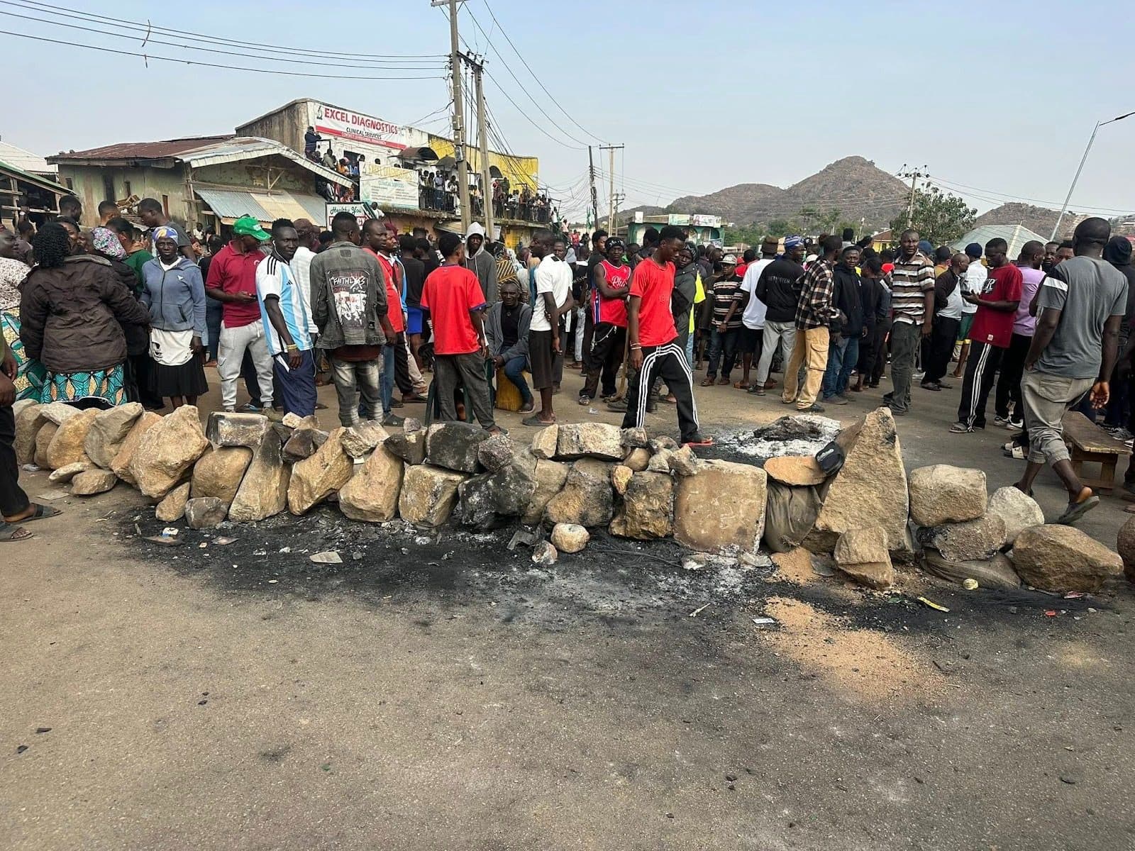A large crowd gathers near a barricade of rocks on a street, with hills visible in the background. Some buildings and power lines are nearby.