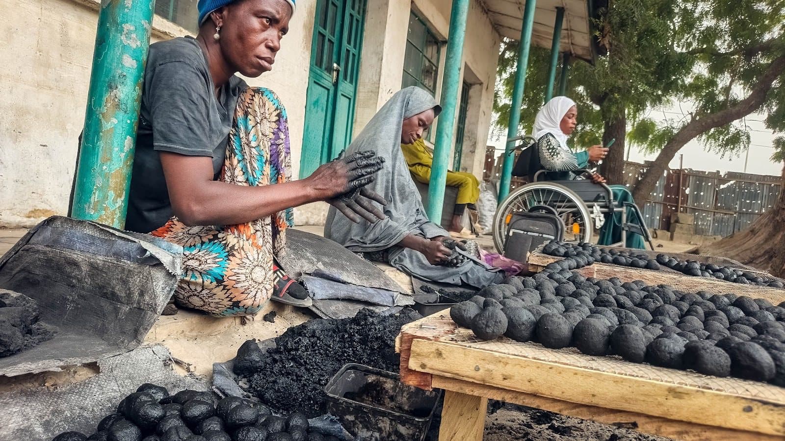 People forming coal balls by hand, sitting outdoors. One person in a wheelchair, another in colorful attire, surrounded by trays of coal balls.