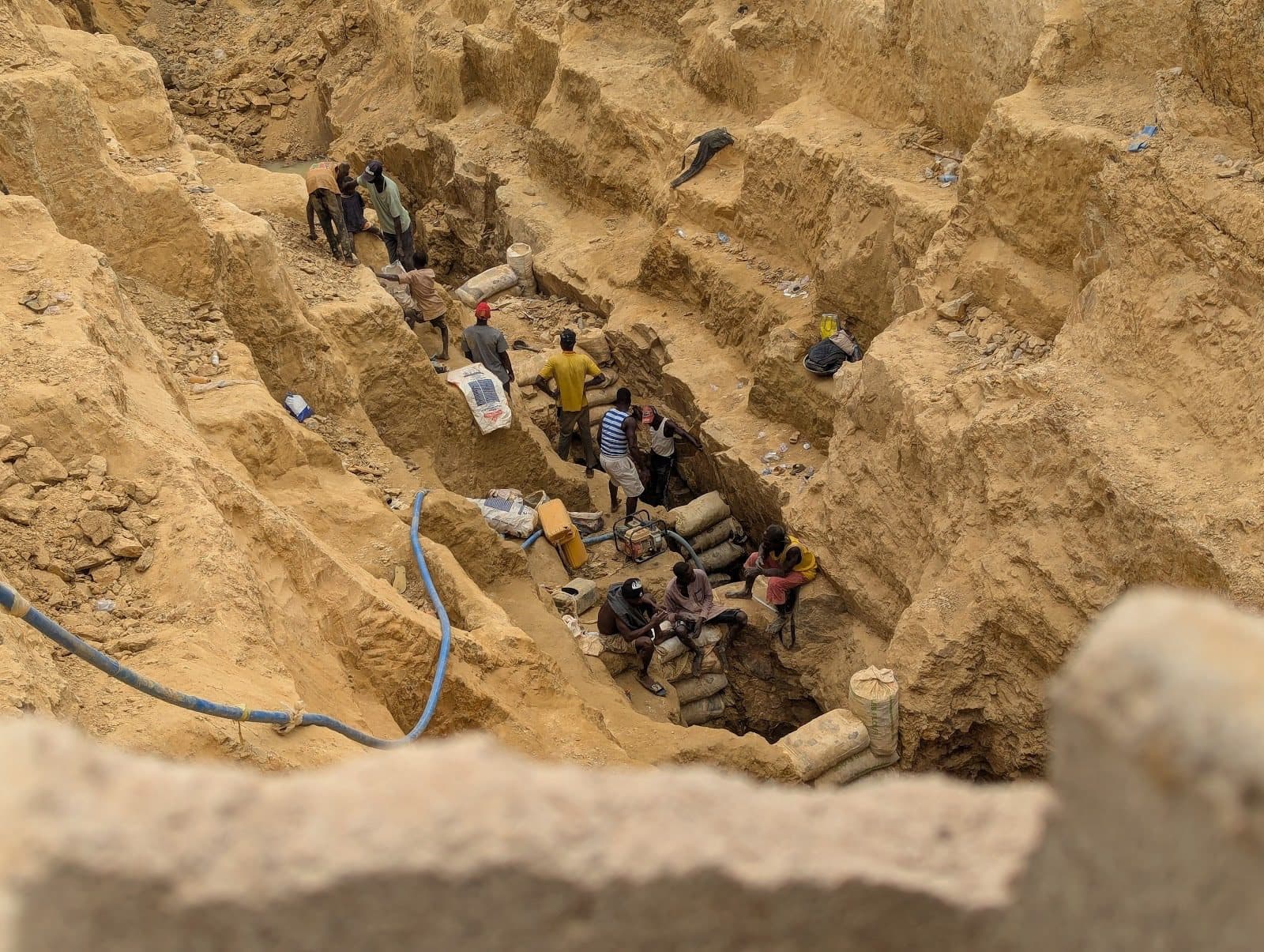 People working in a deep, narrow excavation site with exposed earth walls, carrying bags and using tools.