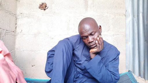 Person in a blue outfit appears deep in thought, sitting against a light-colored wall, with head resting on hand.