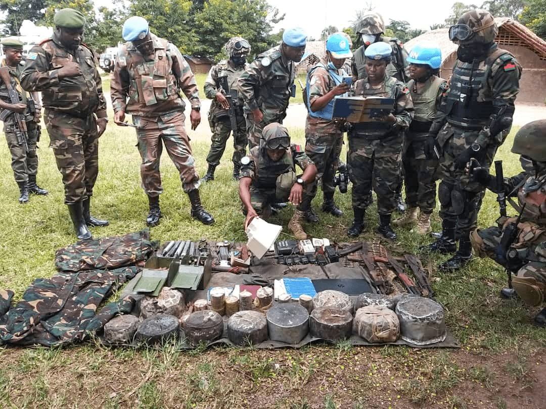Soldiers in camouflage and UN helmets examine items laid out on the ground, discussing and taking notes.