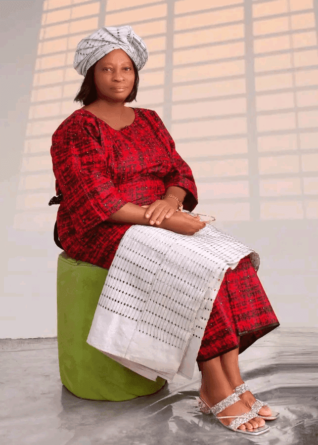A woman in a red traditional outfit with a matching headwrap sits on a green stool, holding a white cloth, indoors.