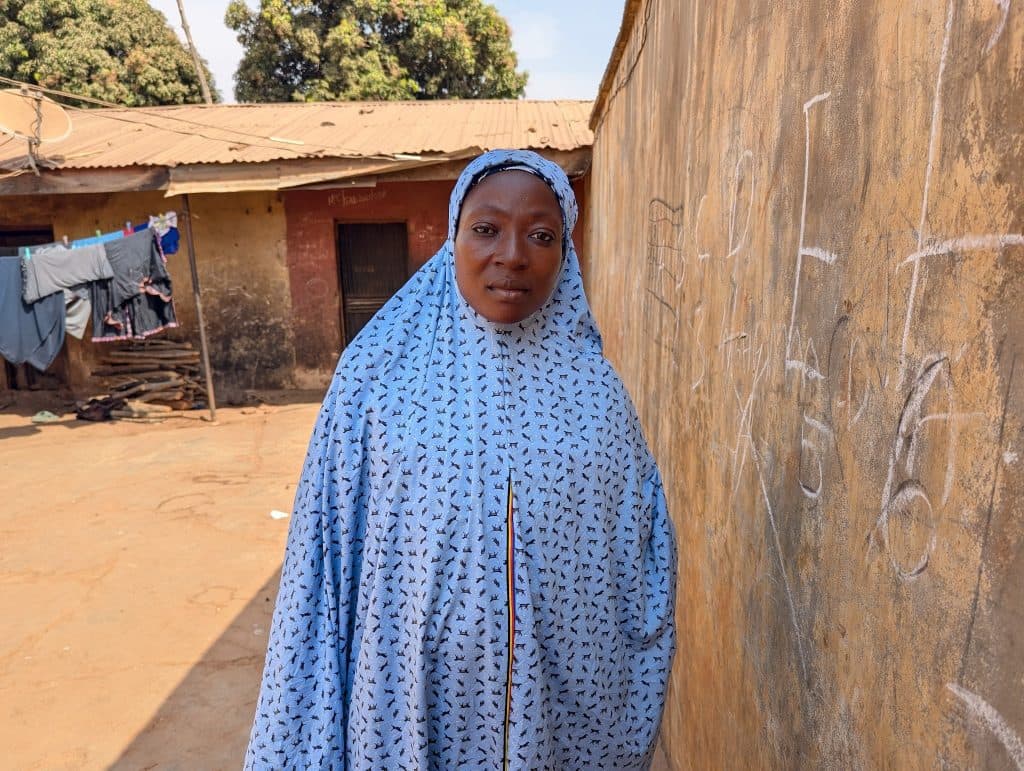 Woman in a light blue patterned hijab stands beside a wall with chalk markings, with laundry hanging in the background.
