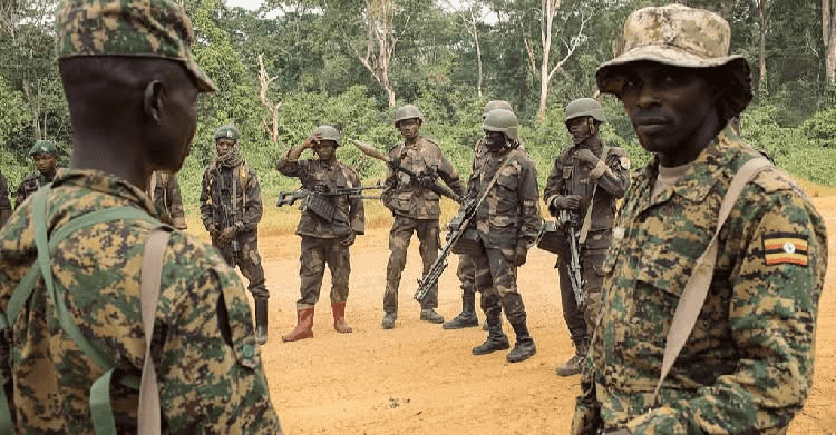Soldiers in camouflage uniforms stand on a dirt path, equipped with rifles and wearing helmets, surrounded by green foliage.
