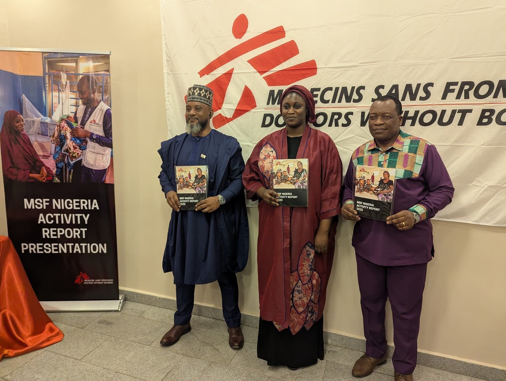 Three people stand holding MSF Nigeria activity reports in front of a banner. A sign reads "MSF Nigeria Activity Report Presentation."