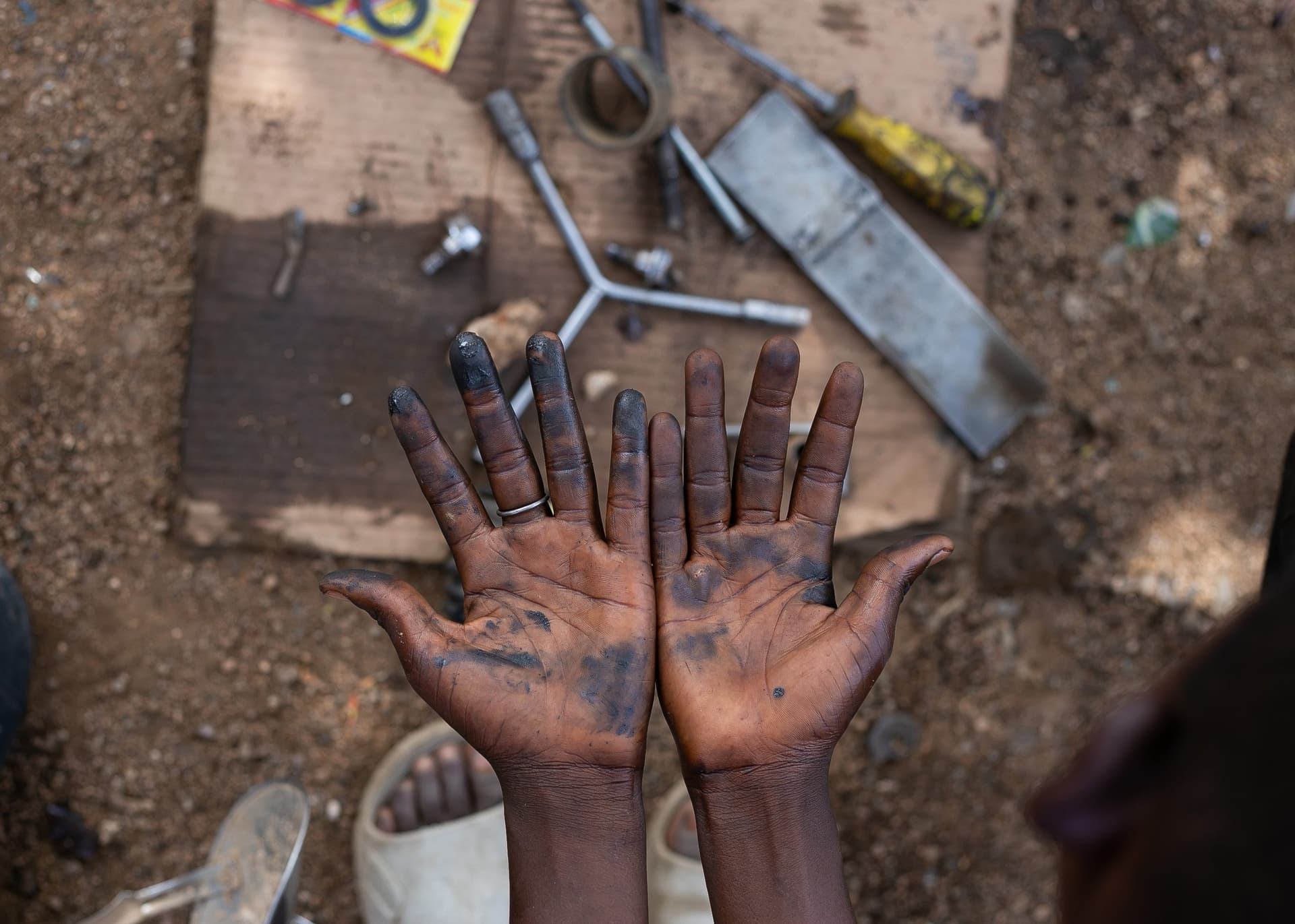 Dirty hands of a mechanic, open palms showing grease, with scattered tools on the ground.