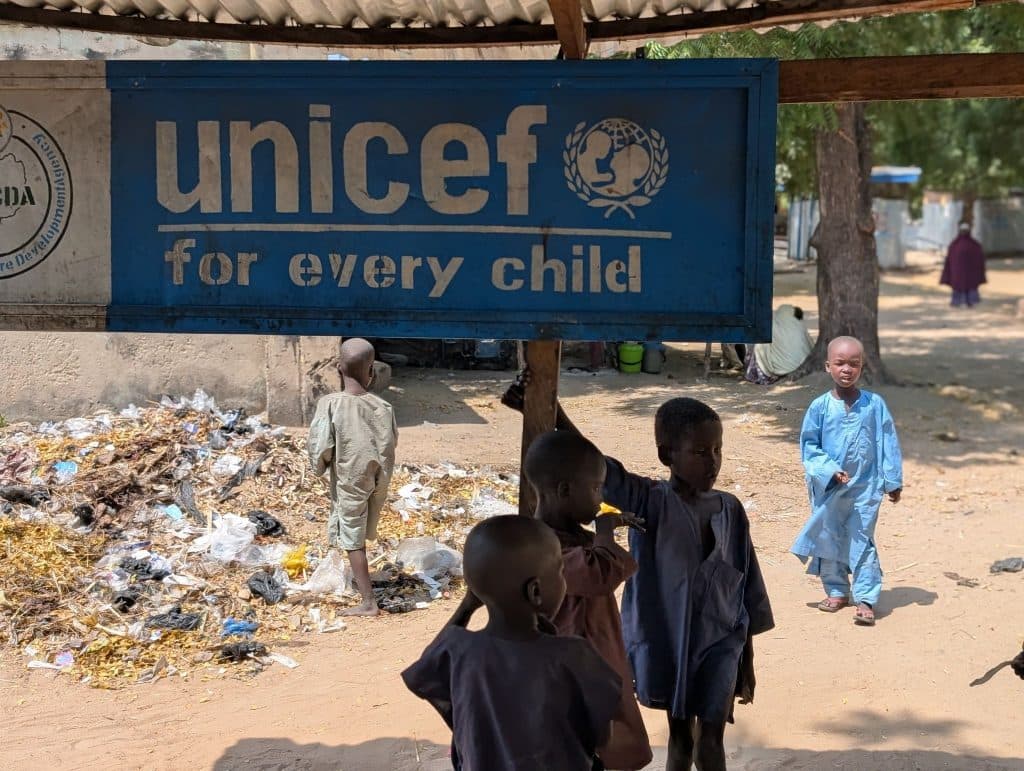 Children underneath a "UNICEF for every child" sign, standing on a dirt ground with scattered litter.