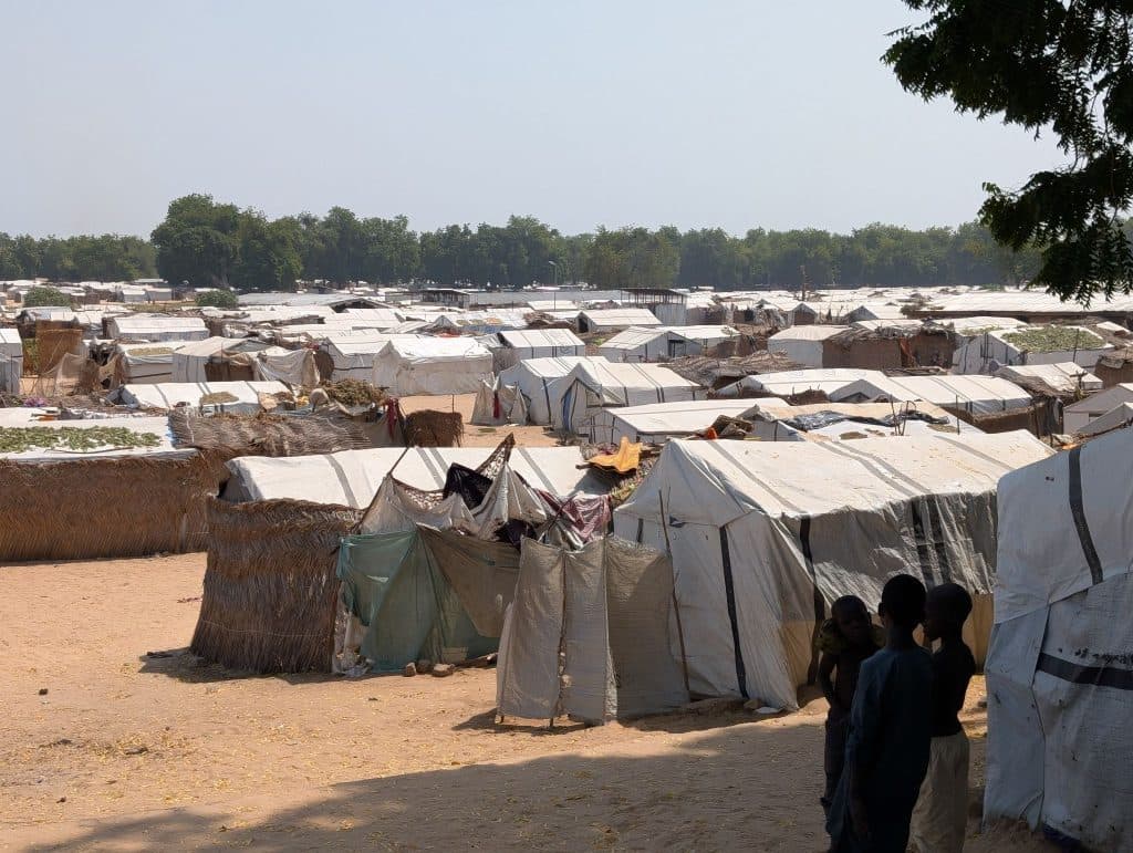 A vast view of a makeshift camp with tents and temporary shelters, three children stand in the foreground, trees line the background.