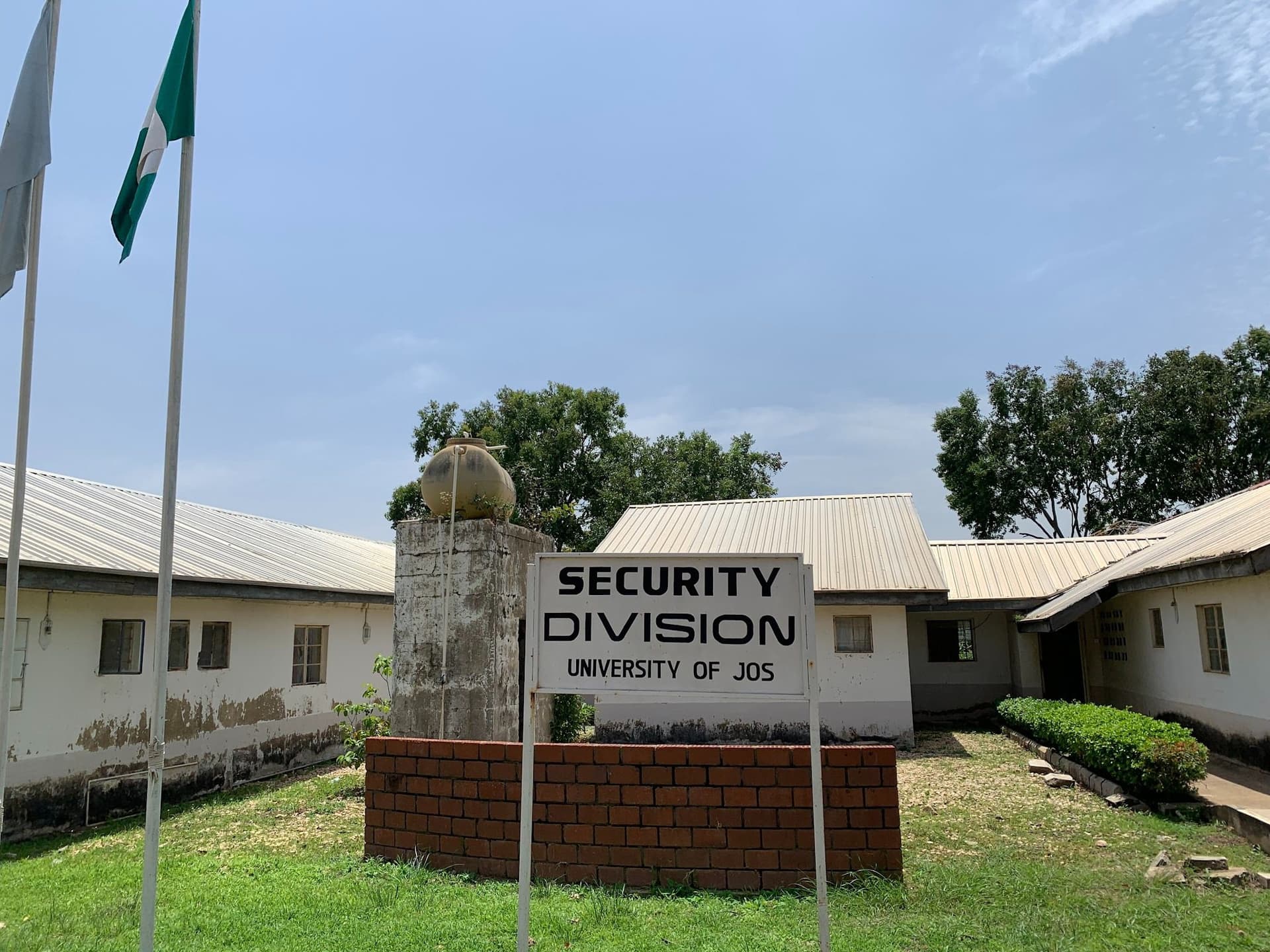 Security Division sign at University of Jos, flanked by flagpoles, with a white building and trees in the background.
