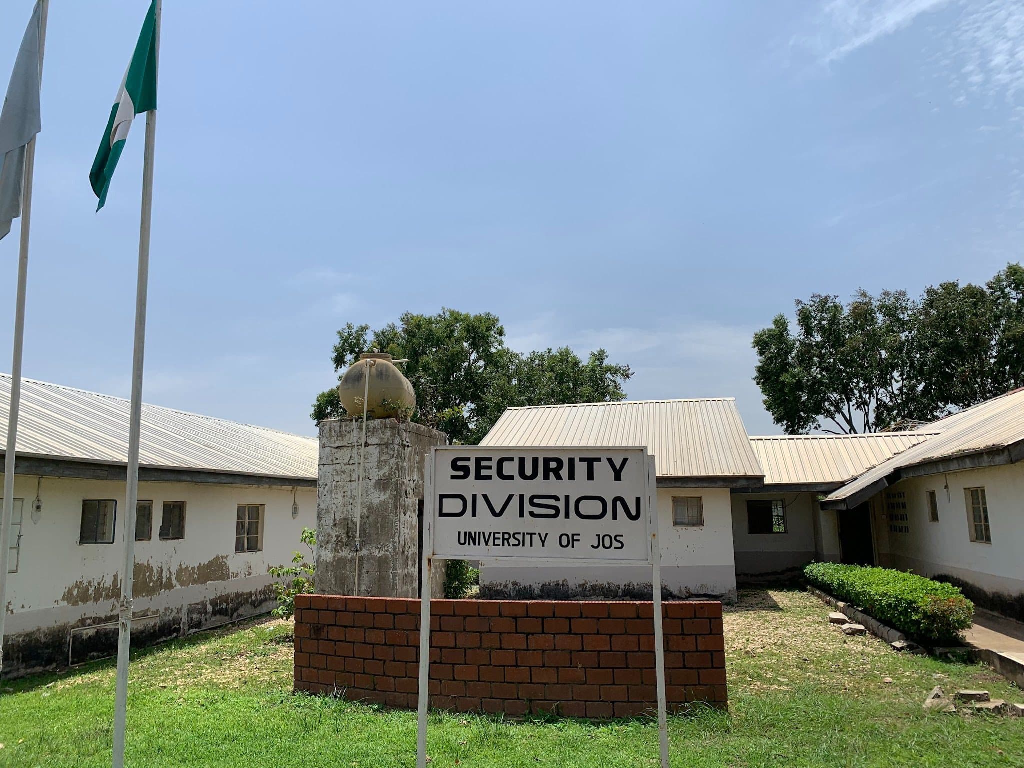 Security Division sign at University of Jos, flanked by flagpoles, with a white building and trees in the background.