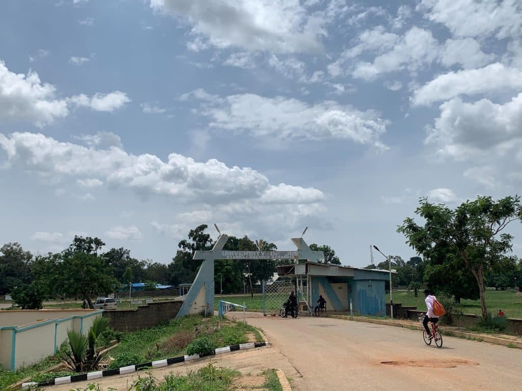 Entrance of a university with people cycling and walking, surrounded by greenery and a partly cloudy sky.