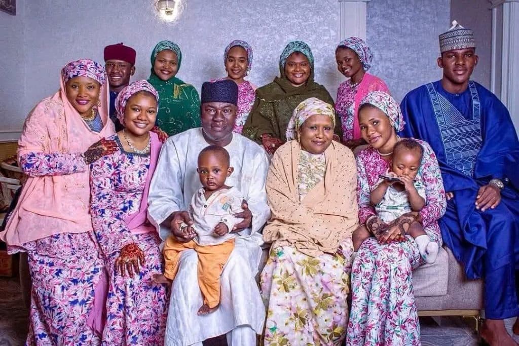 A family of thirteen poses indoors, wearing colorful traditional attire, with two adults holding young children.