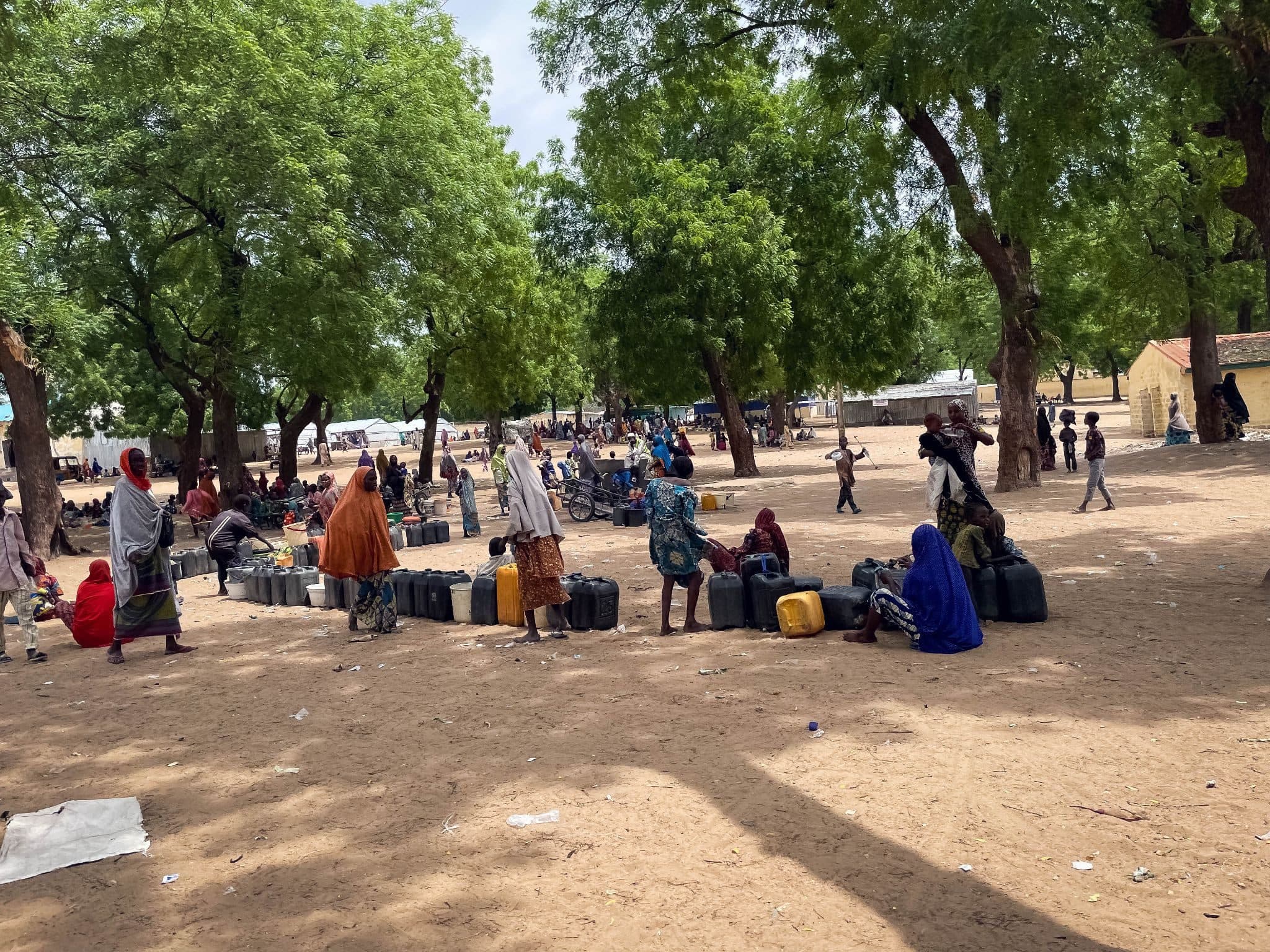 People gather under trees, with containers, in a sandy open area, while others are in the background near buildings.