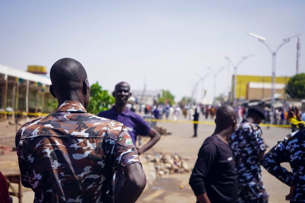People in camouflage uniforms and plainclothes gather on a street, with others in the background past yellow tape.
