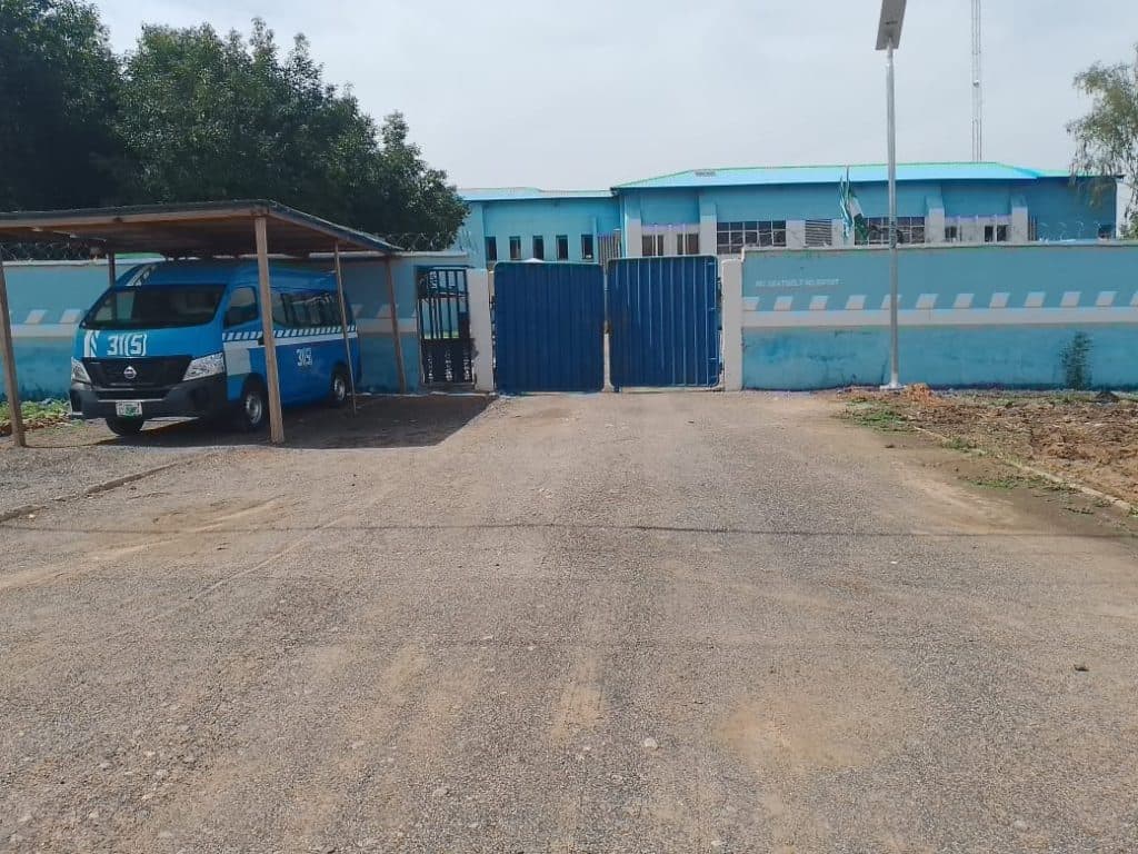 A blue van is parked under a shelter next to a blue gate, with a large blue building in the background.