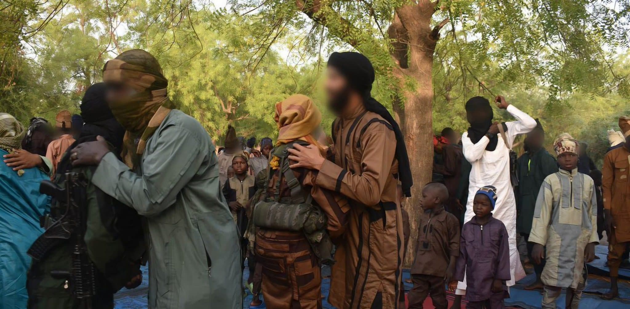A group of people in the outdoors, some wearing camouflage and face coverings, others in traditional attire. Trees are in the background.