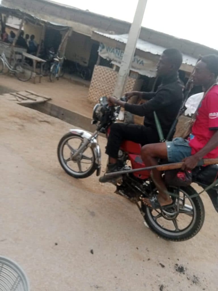 Two people riding a motorcycle on a street, passing by a building with people and bicycles in the background.