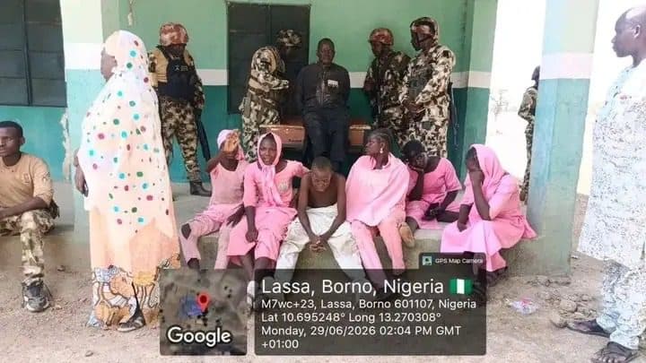 A group of people in traditional attire sit and stand outside a building in Lassa, Borno, Nigeria, alongside military personnel.