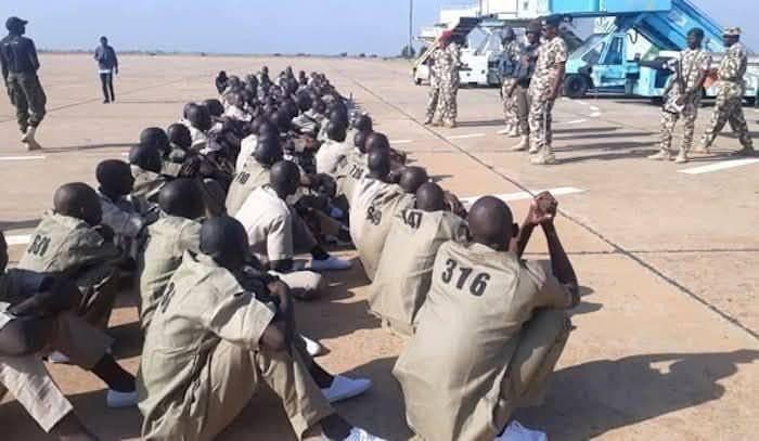 A group of seated individuals in uniformed khaki outfits, numbered on their backs, listen to military personnel on an airfield.