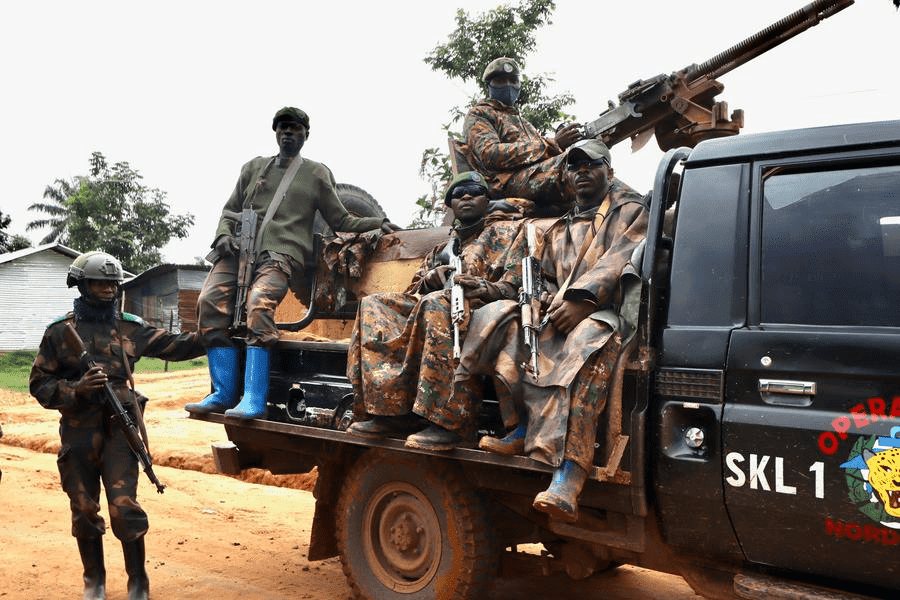 Soldiers in camo sit on a truck mounted with a gun, while another stands nearby with a rifle. Trees and structures in the background.