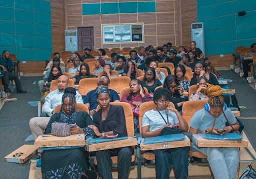A group of students sitting in a lecture hall, some are looking at their phones while others face forward attentively.