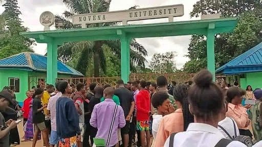 A large group of people gathered at the entrance of Novena University, with a green building in the background.