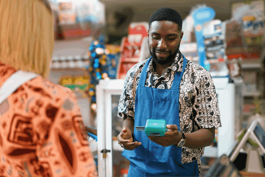 Store clerk in a blue apron processing a payment with a card reader while interacting with a customer at the counter.