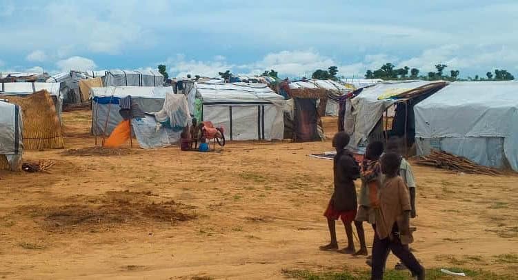 Children walk near makeshift tents in a dry, open area under a cloudy sky.