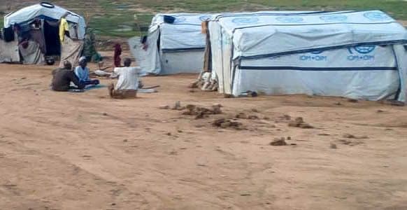 Tents on a dirt path with people sitting outside on the ground, and laundry hanging nearby.