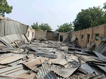 Debris of a collapsed building with corrugated metal sheets scattered on the ground under a clear sky. Trees are visible in the background.