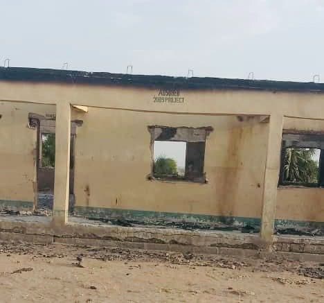 An abandoned, partially burnt building with missing windows and a damaged roof, surrounded by a barren landscape.