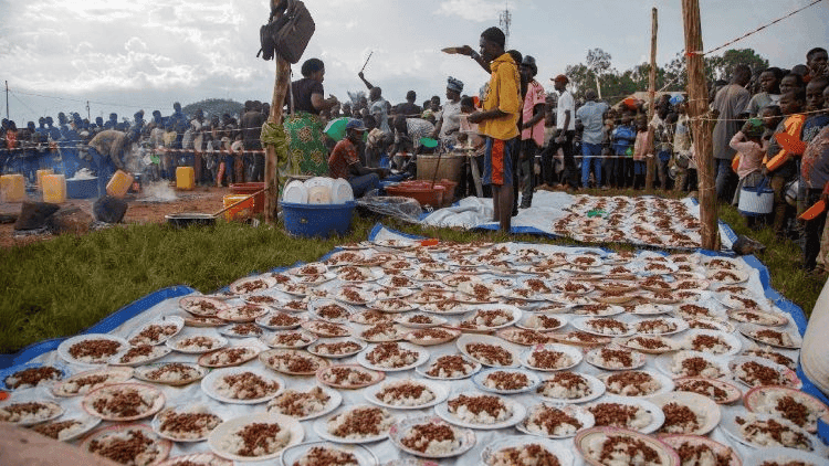 Plates of food arranged on a tarp while a crowd gathers at an outdoor event.