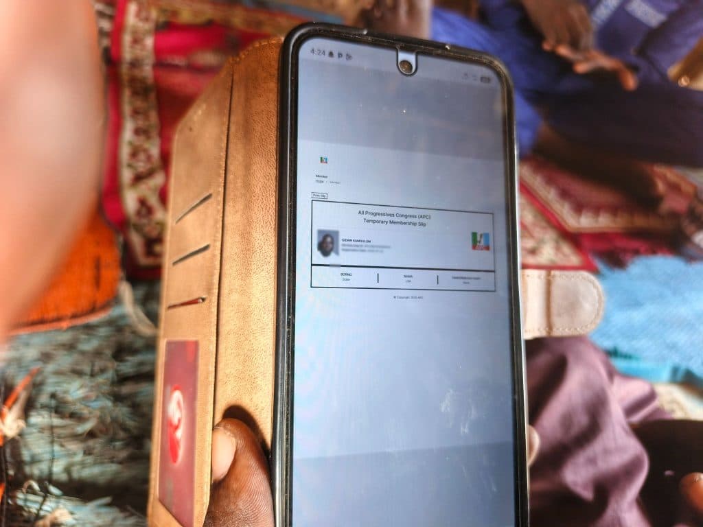 Person holding a smartphone displaying an All Progressives Congress (APC) temporary membership slip, with colorful fabrics in the background.