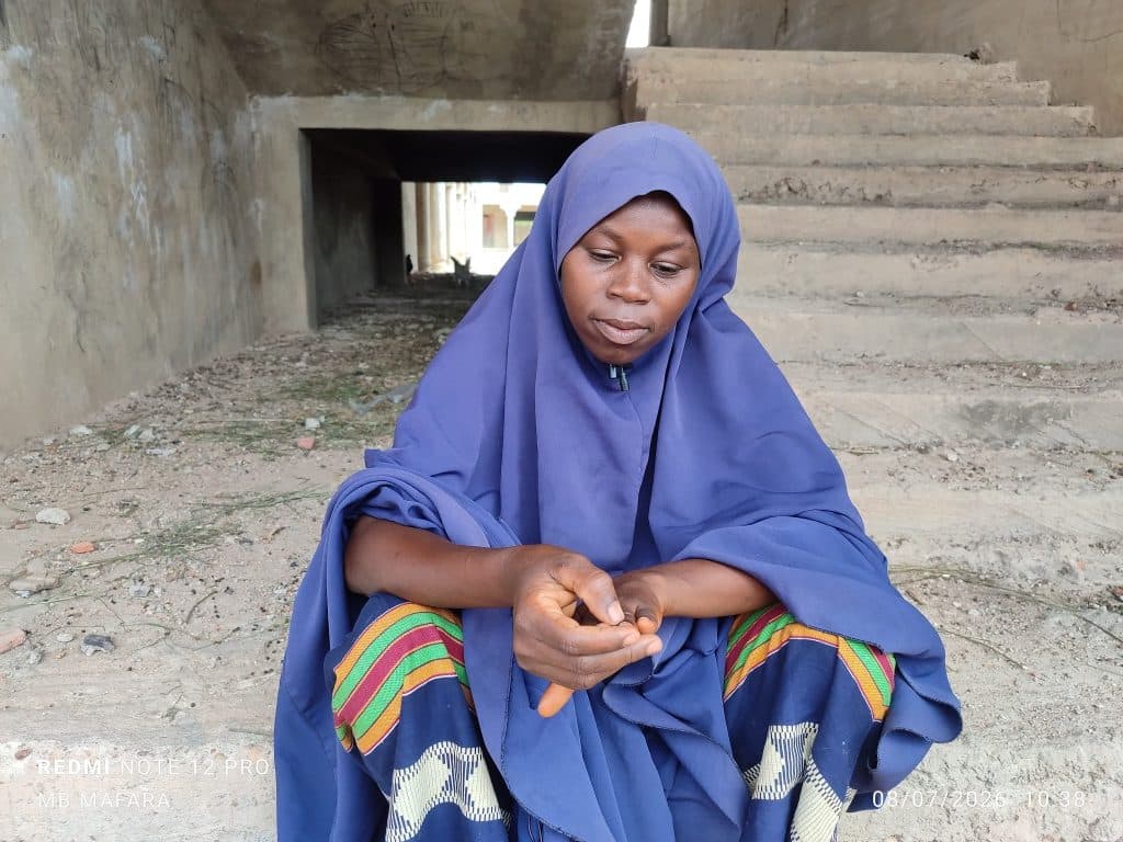 A woman in a blue headscarf sits on a step under a concrete structure, looking down thoughtfully.