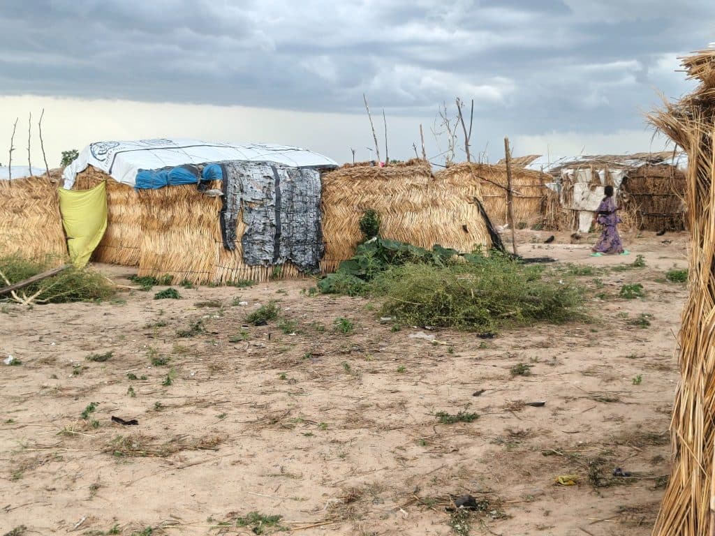Straw huts with makeshift covers under a cloudy sky, a person walks by in the background. Sparse vegetation on sandy ground.