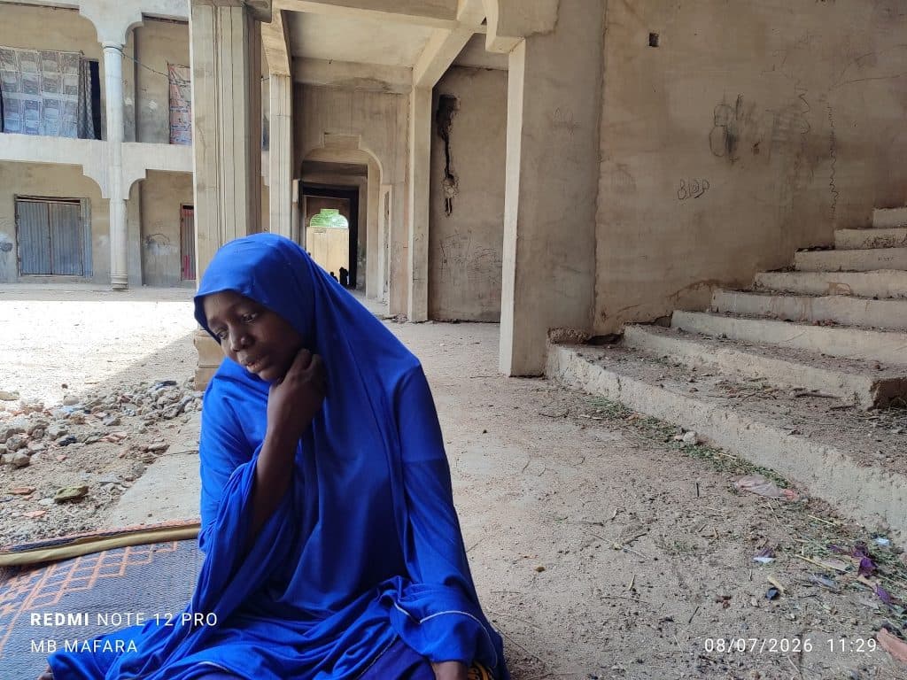 Woman in a blue hijab sits in a partially constructed building with concrete columns and stair steps.